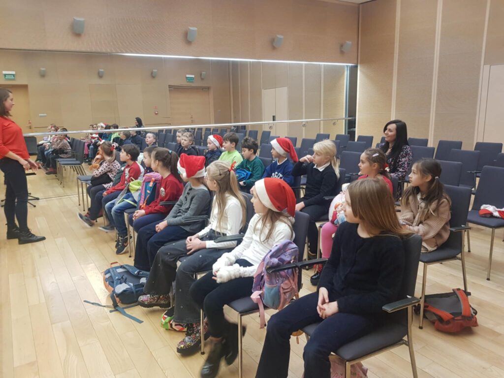 W kameralnej sali koncertowej, na krzesłach siedzi grupa dzieci. Zajmują dwa rzędy. Z przodu stoi kobietra w czerwonym swetrze. Mówi do publiczności.