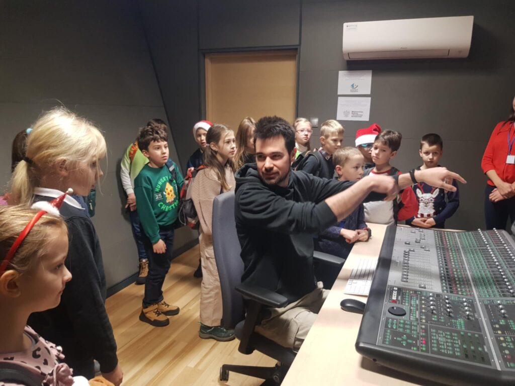 W niewielkim pomieszczeniu centralne miejsce zajmuje konsola z wieloma przyciskami i suwakami. Przed nią siedzi młody mężczyzna odwrócony do tyłu, patrzy w strone dzieci, które stoją za nim, pod ścianą.