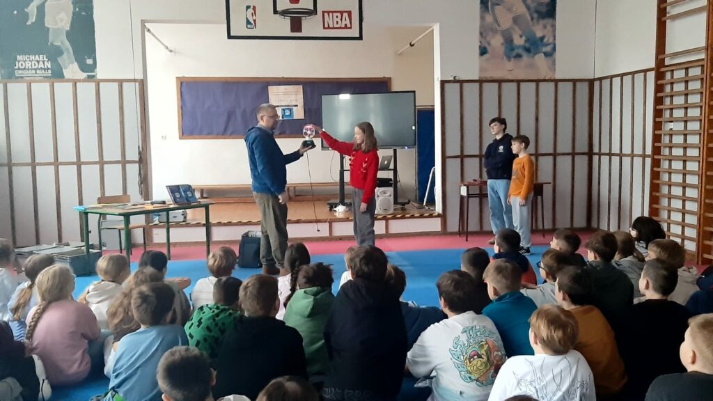 Zdjęcie przedstawia mężczyznę trzymającego kulę plazmową przed nim dziewczynka na kuli kładzie dłoń. W oddali stoi dwóch chłopców przyglądają się doświadczeniu Za nimi widać ekran, tablicę na ścianie z granatowym tłem na której widać powieszony plakat, obok mężczyzny jest stolik z książkami. Przed osobami stojącymi siedzą widzowie tyłem do fotografa.