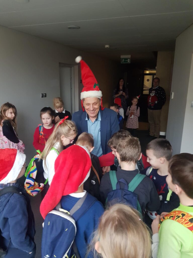 Na środku pomieszczenia stoi uśmiechniety mężczyzna, w czapce Mikołaja. Rozdaje cukierki dzieciom, które stoja przed nim.