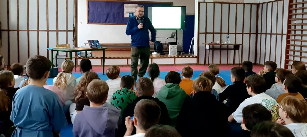 Zdjęcie przedstawia mężczyznę z mikrofonem w dłoni przemawiającego do dużej grupy dzieci siedzących przed nim.