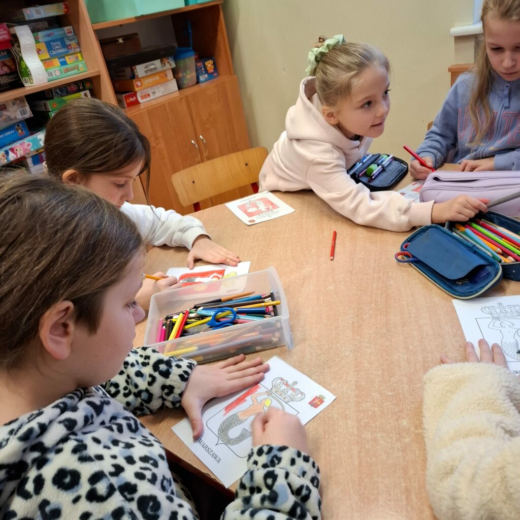 Zdjęcie pokazuje cztery dziewczynki siedzące przy brązowym sześciokątnym stole. Dziewczynki kolorują kredkami herb Warszawy. Jedna z dziewczynek wyciąga rękę w stronę piórnika. W tle kremowa ściana oraz brązowa szafka z kolorowymi zabawkami.