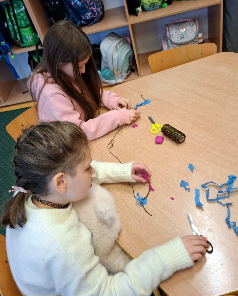 Zdjęcie przedstawia dwie dziewczynki siedzące przy brązowym sześciokątnym stole. W rękach mają kolorowe bibułki i nożyczki. W tle szafki z tornistrami.