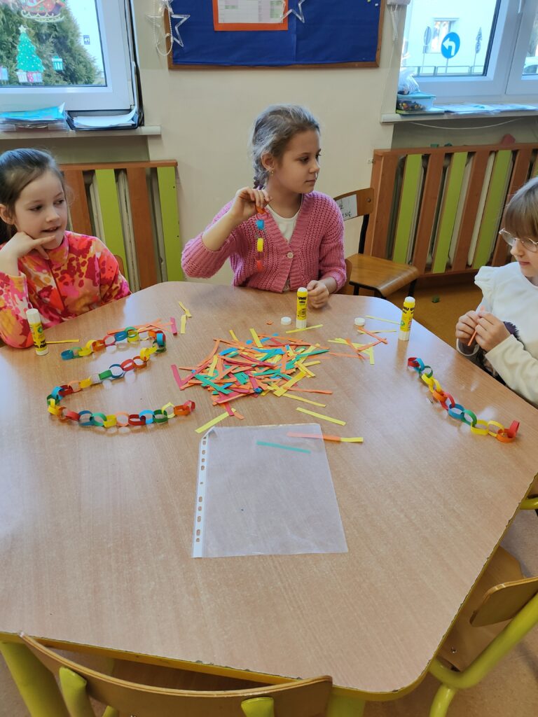 Zdjęcie przedstawia trzy dziewczynki siedzące przy brązowym sześciokątnym stole. W rękach mają kolorowe paski papieru. Na stole leżą kleje, kolorowe paski papieru oraz kolorowy łańcuch z papieru. W tle ściana, kaloryfer i dwa okna.