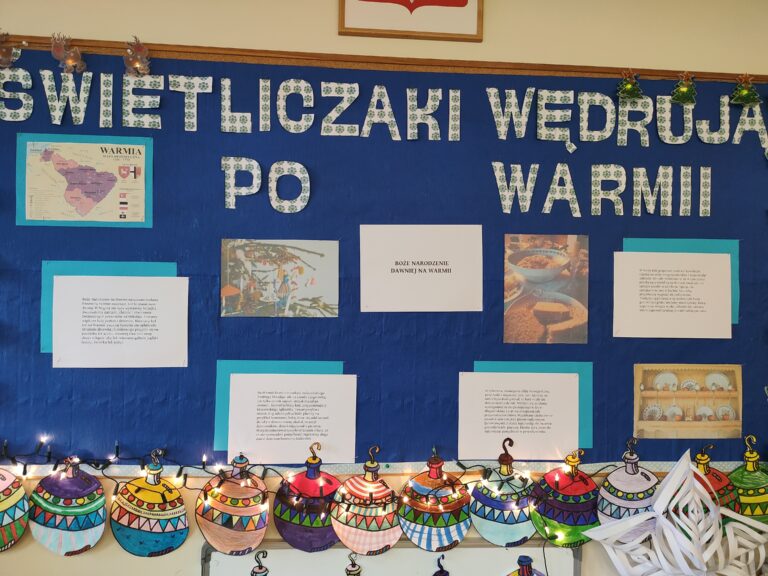 Zdjęcie przedstawia gazetkę szkolną wiszącą na kremowej ścianie. Tło jest granatowe, na nim napis Świetliczaki wędrują po Warmii” . Są także zdjęcia oraz kartki z napisami. Na dole przyczepione są kolorowe kolorowanki przedstawiające piękne bombki.