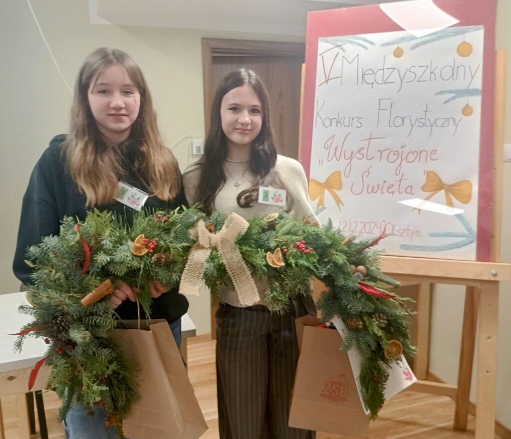 Na zdjęciu widzimy dwie stojące dziewczynki, które trzymają girlandę świąteczną. Girlanda wykonana została z naturalnych gałązek jodły i tui. Przyozdobiona została suszonymi plastrami pomarańczy, szyszkami, cynamonem, szyszkami oraz kulkami żurawiny i jarzębiny. Centralnym elementem girlandy jest kokarda z naturalnego lnu, która dodaje całości elegancji i rustykalnego charakteru. Dziewczynki trzymają również torby papierowe, w których najprawdopodobniej umieszczone są nagrody. Obok widoczna jest tablica z napisem „V Międzyszkolny Konkurs Florystyczny” oraz hasłem „Wystrojone Święta”, które nawiązują do tematyki świątecznej.