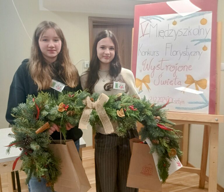Na zdjęciu widzimy dwie stojące dziewczynki, które trzymają girlandę świąteczną. Girlanda wykonana została z naturalnych gałązek jodły i tui. Przyozdobiona została suszonymi plastrami pomarańczy, szyszkami, cynamonem, szyszkami oraz kulkami żurawiny i jarzębiny. Centralnym elementem girlandy jest kokarda z naturalnego lnu, która dodaje całości elegancji i rustykalnego charakteru. Dziewczynki trzymają również torby papierowe, w których najprawdopodobniej umieszczone są nagrody. Obok widoczna jest tablica z napisem „V Międzyszkolny Konkurs Florystyczny” oraz hasłem „Wystrojone Święta”, które nawiązują do tematyki świątecznej.