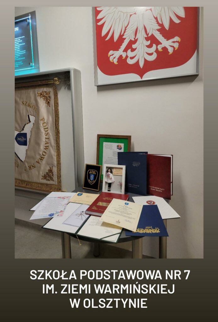 Zdjęcie przedstawia wystawę nagród i wyróżnień w Szkole Podstawowej nr 7 im. Ziemi Warmińskiej w Olsztynie. Na stole znajdują się różne dyplomy, podziękowania, dokumenty i odznaczenia, świadczące o osiągnięciach placówki. W tle widoczny jest fragment sztandaru szkoły z jej nazwą oraz biały orzeł w czerwonym polu – godło Polski. Całość prezentuje uroczystą i oficjalną atmosferę, podkreślającą wagę prezentowanych dokumentów i związane z nimi sukcesy.