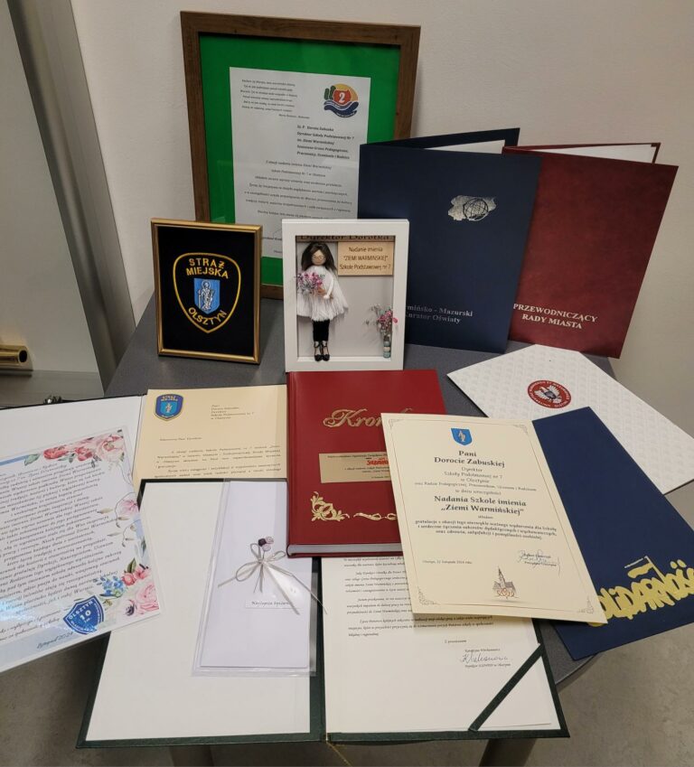 Zdjęcie przedstawia zbiór wyróżnień, dyplomów i podziękowań wyeksponowanych na stole. W centralnej części widoczne są elegancko oprawione dokumenty, takie jak dyplomy, pisma z herbem oraz ozdobny akt nadania szkole imienia „Ziemi Warmińskiej”. Wśród przedmiotów znajdują się również: Tabliczka z herbem Straży Miejskiej w Olsztynie. Ramka z figurką dziewczynki w stroju regionalnym oraz dedykacją nawiązującą do nadania imienia szkole. Dokumenty w ciemnych teczkach oznaczonych symbolami instytucji, takich jak Rada Miasta czy Kuratorium Oświaty. Listy i podziękowania ozdobione grafikami, w tym kwiatami, które podkreślają uroczysty charakter tych materiałów. Całość tworzy elegancką kompozycję, która podkreśla wyjątkowe wydarzenia w życiu szkoły i jej współpracę z różnymi instytucjami.