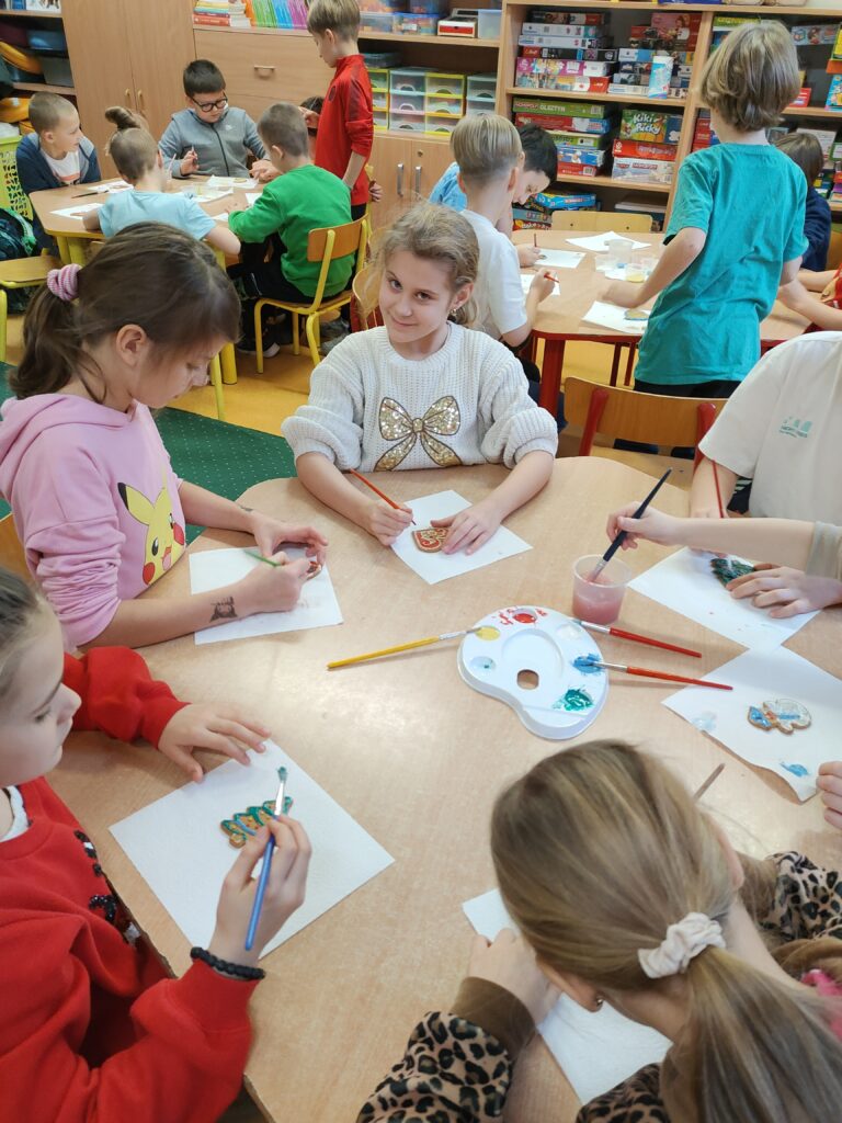 Na zdjęciu widzimy grupę dzieci siedzących przy stolikach w sali szkolnej i zajmujących się dekorowaniem pierników lub podobnych przedmiotów. Każde dziecko jest skupione na swojej pracy, korzystając z pędzli, farb i innych przyborów plastycznych. W tle widać regały z grami, książkami i materiałami edukacyjnymi, co podkreśla kreatywny charakter zajęć. Atmosfera jest przyjazna i twórcza, a dzieci wydają się zadowolone z udziału w tej aktywności.