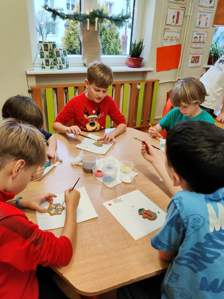 Na zdjęciu widać grupę chłopców siedzących przy stoliku i malujących ozdobne pierniki. W tle znajduje się okno udekorowane świątecznym girlandem co nadaje pomieszczeniu świątecznego klimatu. Na stole leżą farby, pędzle i gotowe pierniki, nad którymi dzieci pracują z dużym skupieniem. Atmosfera jest ciepła i kreatywna, a dzieci wydają się cieszyć wspólną pracą twórczą.