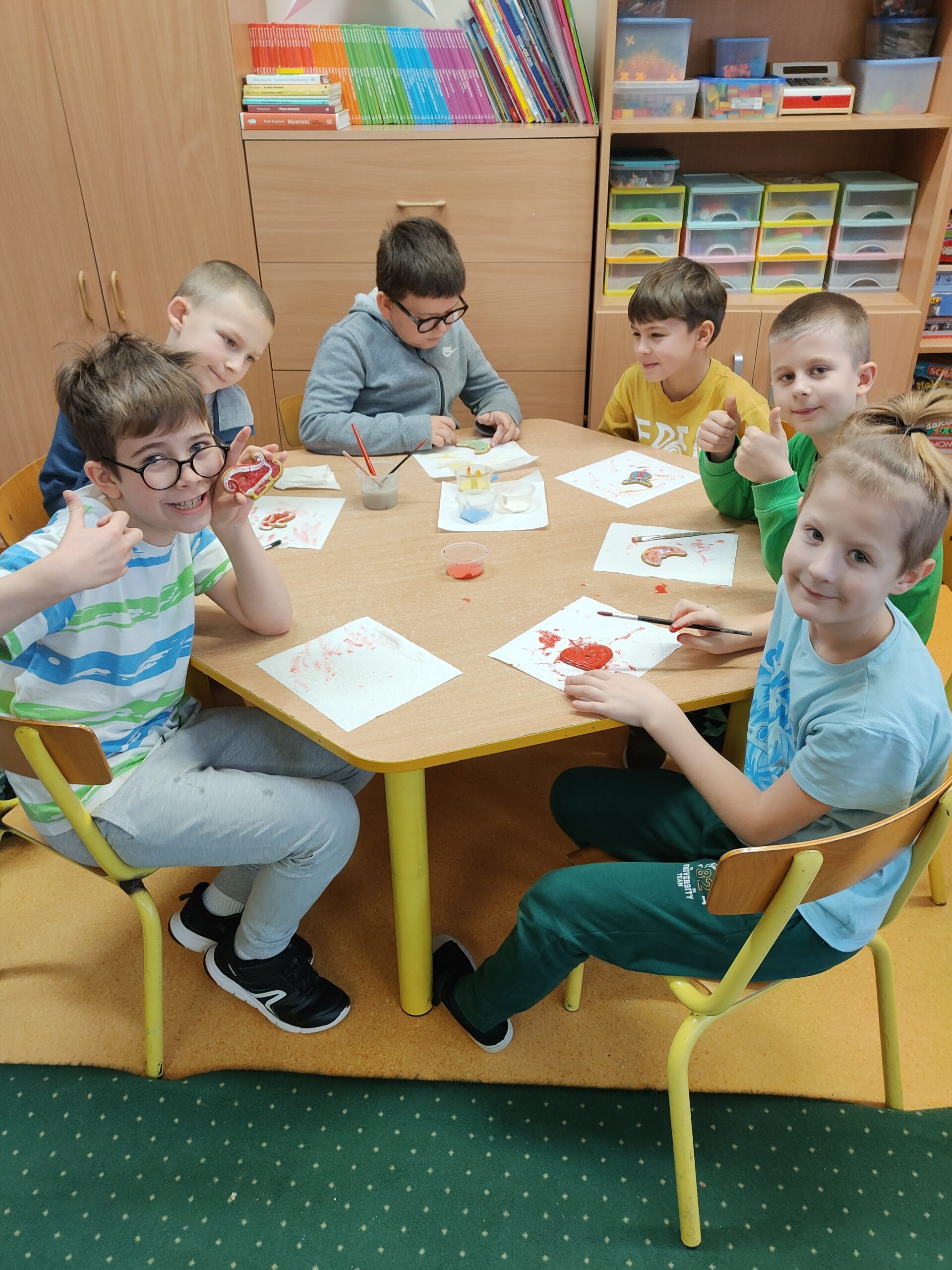 Na zdjęciu znajduje się grupa sześciu dzieci siedzących przy stole w sali szkolnej lub przedszkolnej. Dzieci zajmują się pracami plastycznymi, malując lub dekorując różne elementy. W tle widać półki z książkami, kolorowymi materiałami i pojemnikami z przyborami do zajęć. Atmosfera na zdjęciu jest wesoła, a dzieci wydają się być zaangażowane i uśmiechnięte.