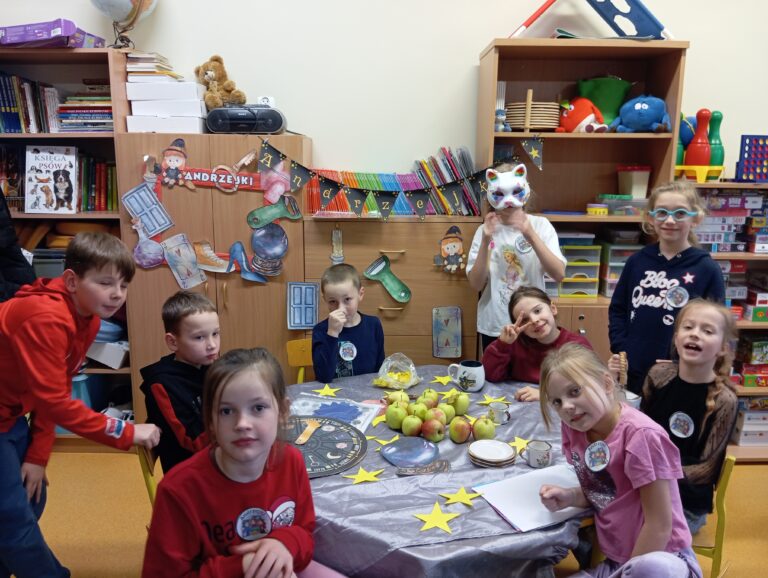 Na zdjęciu widzimy grupę dzieci w sali szkolnej. Na środku stołu leżą jabłka, papierowe gwiazdki oraz inne przedmioty, które mogą być związane z wróżbami lub zabawą andrzejkową. W tle znajduje się dekoracja z napisem "Andrzejki", przedstawiająca symbole związane z wróżbami, takie jak kryształowa kula i karty.