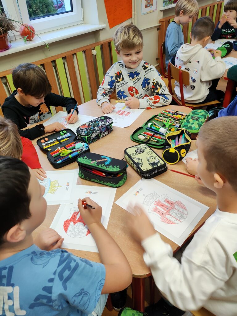 Zdjęcie przedstawia grupę chłopców siedzących za sześciokątnym, brązowym stołem. Są oni ubrani w kolorowe ubrania. Kolorują karty pracy z herbem Krakowa. Na stole leżą kolorowanki oraz kolorowe piórniki. W tle widać kremową ścianę i kaloryfer z zielono – brązową zabudową. Widać także okno, biały parapet, na którym stoi w brązowej doniczce zielony kwiat oraz leży pomarańczowa dynia. W oddali widać inny stolik, przy który siedzi trójka chłopców.