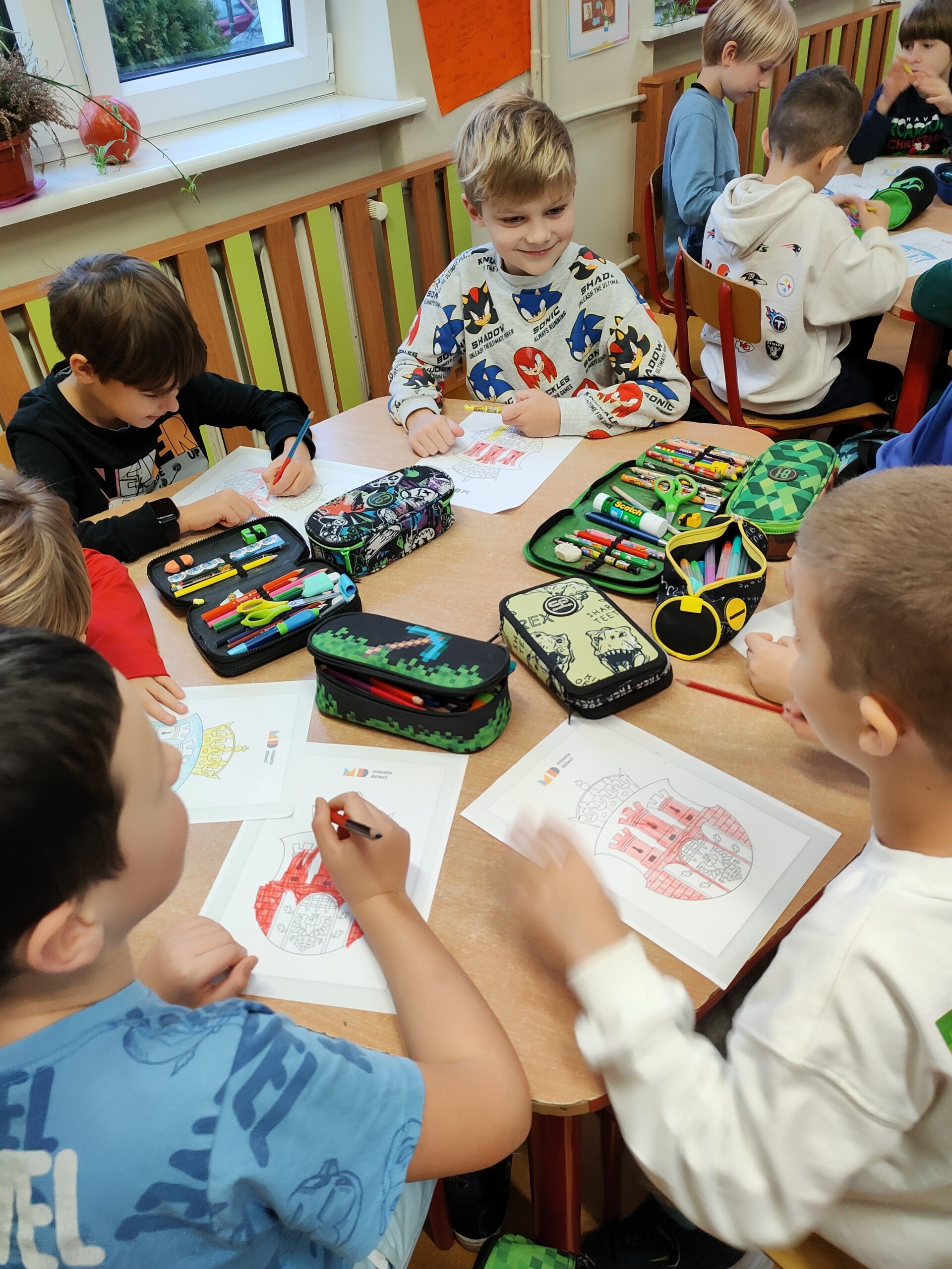 Zdjęcie przedstawia grupę chłopców siedzących za sześciokątnym, brązowym stołem. Są oni ubrani w kolorowe ubrania. Kolorują karty pracy z herbem Krakowa. Na stole leżą kolorowanki oraz kolorowe piórniki. W tle widać kremową ścianę i kaloryfer z zielono – brązową zabudową. Widać także okno, biały parapet, na którym stoi w brązowej doniczce zielony kwiat oraz leży pomarańczowa dynia. W oddali widać inny stolik, przy który siedzi trójka chłopców.