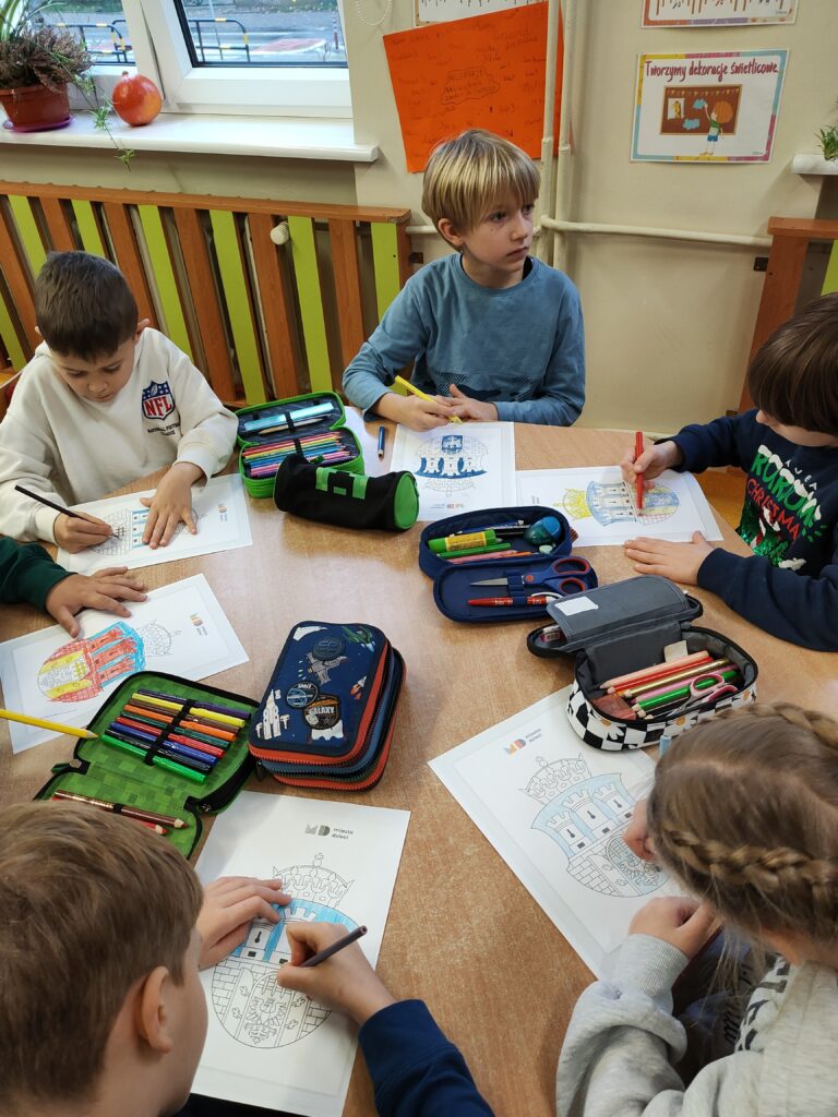 Zdjęcie przedstawia czterech chłopców oraz jedną dziewczynkę, siedzących za sześciokątnym, brązowym stołem. Dzieci ubrane są w kolorowe ubrania. Kolorują karty pracy z herbem Krakowa. Na stole leżą kolorowanki oraz kolorowe piórniki. W tle widać kremową ścianę i kaloryfer z zielono – brązową zabudową. Widać także okno, biały parapet, na którym stoi w brązowej doniczce zielony kwiat a także lezy pomarańczowa dynia.