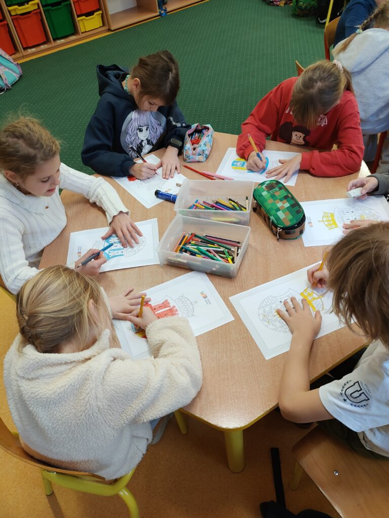 Zdjęcie przedstawia czterech chłopców oraz jedną dziewczynkę, siedzących za sześciokątnym, brązowym stołem. Dzieci ubrane są w kolorowe ubrania. Kolorują karty pracy z herbem Krakowa. Na stole leżą kolorowanki oraz kolorowe piórniki. W tle widać kremową ścianę i kaloryfer z zielono – brązową zabudową. Widać także okno, biały parapet, na którym stoi w brązowej doniczce zielony kwiat a także lezy pomarańczowa dynia.