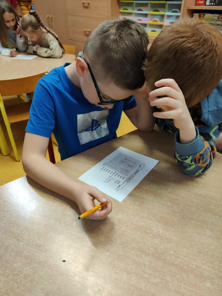 Zdjęcie przedstawia siedzących przy brązowym , sześciokątnym stole chłopców. Pierwszy z nich ubrany jest w granatową koszulkę, na nosie ma granatowe okulary. W ręku trzyma żółty ołówek. Pochyla się nad kartą pracy, jaką jest wykreślanka. Drugi chłopiec, który pochyla się nad kartką ma rude włosy. Widać dłoń i kawałek kolorowej bluzy. W oddali widzimy inny stolik i siedzące przy nim dwie dziewczynki. W tle widzimy brązowe szafki i kolorowe pudełeczka.