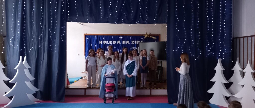 Dzieci wraz z nauczycielką uczestniczą w wydarzeniu szkolnym, gdzie śpiewają kolędę w ramach Szkolnego klasowego kolędowania. Na zdjęciu grupa uczniów stojących na poziomie podwyższenia. W tle znajduje się dekoracja z niebieską tkaniną, na której znajdują się napisy „KOLĘDA NA ZIMĘ” białe gwiazdki i złota kometa. Na pierwszym planie, po prawej stronie, stoi wychowawczyni ubrana w jasną bluzkę i spódnicę. Dzieci są ubrane w jasnych strojach – niektóre w białych, przypominających anioły, inne w odświętnych ubraniach. Jedna z osób, symbolizująca postać biblijną, trzymająca wózek dziecięcy jako rekwizyt. Scena jest ozdobiona białymi sylwetkami choinek i girland z migoczącymi światłami.