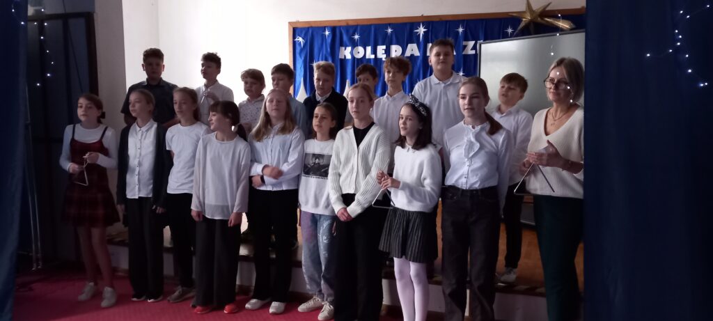 Na zdjęciu grupa chłopców stojąca na poziomie podwyższenia. Stopień niżej ustawione dziewczynki. Wszyscy elegancko ubrani. Niektórzy trzymają trójkąt muzyczny. Na niższej scenie towarzyszy uczniom wychowawczyni. W tle znajduje się dekoracja z niebieską tkaniną, na której znajdują się napisy „KOLĘDA NA ZIMĘ” białe gwiazdki i złota kometa. Po prawej i lewej stronie wisi granatowa kurtyna z girlandą z migoczącymi światłami. Dzieci wraz z nauczycielką uczestniczą w wydarzeniu szkolnym, gdzie śpiewają kolędę w ramach Szkolnego klasowego kolędowania.