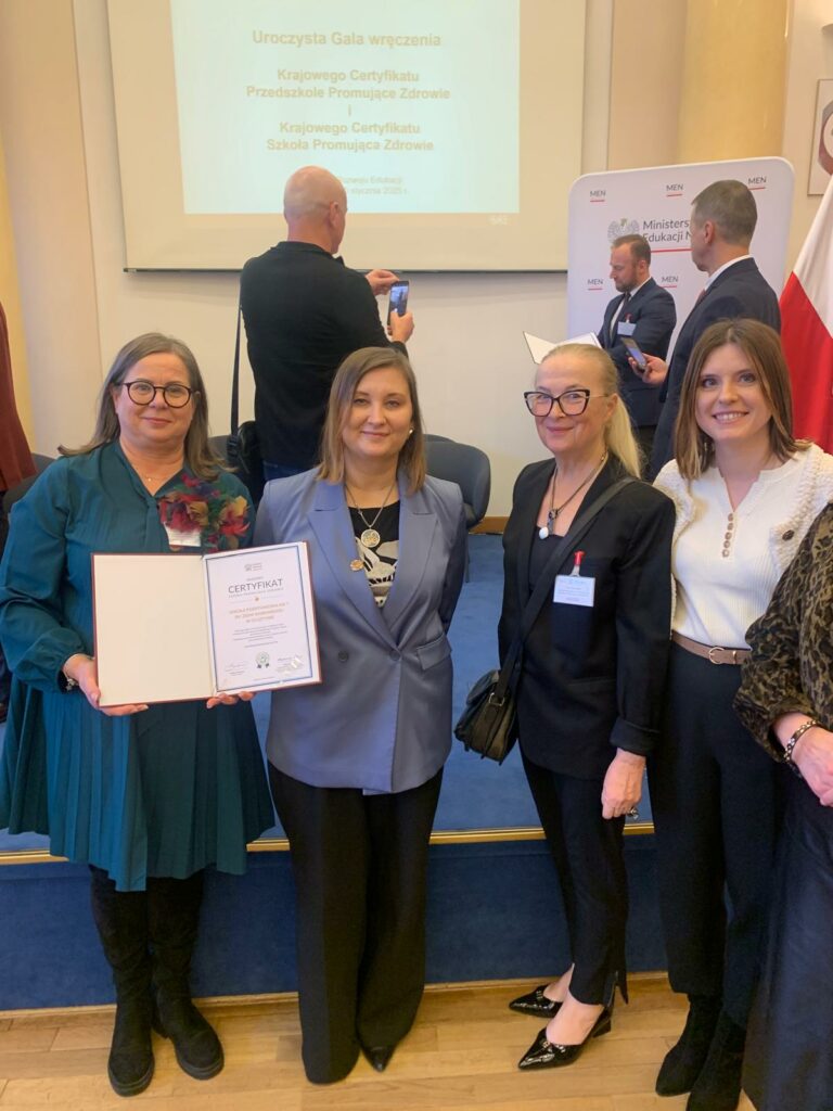 Na zdjęciu znajdują się od lewej strony – Pani Dyrektor Dorota Zabuska trzymająca w dłoniach Krajowy Certyfikat Szkoły Promującej Zdrowie, obok stoi Pełnomocnik Ministra Edukacji do spraw zdrowia psychicznego uczniów - Paulina Piechna-Więckiewicz, kolejno wojewódzki koordynator Kuratorium Oświaty w Olsztynie – Anna Zdaniukiewicz i szkolny koordynator – Diana Gwiazda. W tle znajdują się inne osoby oraz wyświetlany napis – uroczysta gala wręczenia Krajowego Certyfikatu Przedszkole Promujące Zdrowie i Szkoła Promująca Zdrowie.