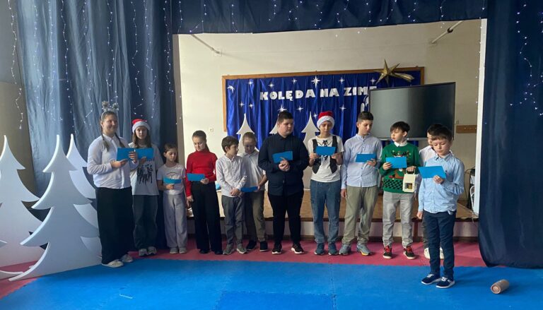 Na zdjęciu grupa 12 uczniów stojących na na scenie. W tle znajduje się dekoracja z niebieską tkaniną, na której znajdują się napisy „KOLĘDA NA ZIMĘ” białe gwiazdki i złota kometa. Wokół nich scenografia z białymi sylwetkami choinek (po lewej stronie) oraz granatowa kurtyna a na niej girlanda z migoczącymi światłami. To uczniowie z Białorusi i Ukrainy oraz przedstawiciele uczniów z Polski, którzy złożyli świąteczne życzenia w swoich ojczystych językach.