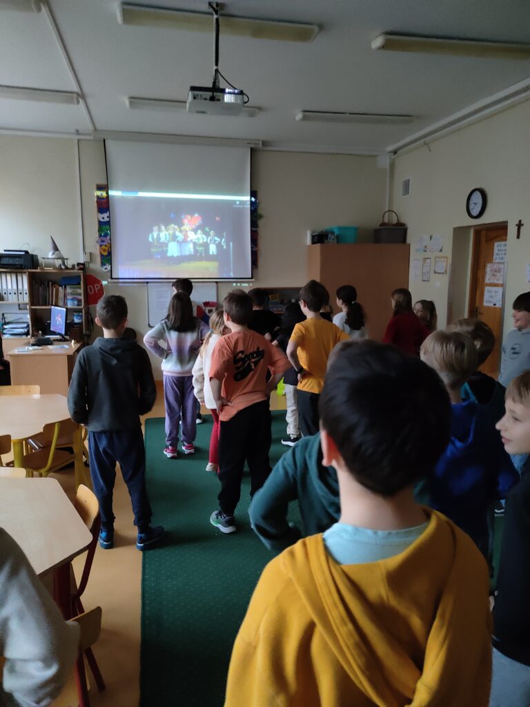 Na zdjęciu widać grupę dzieci stojących w sali lekcyjnej i patrzących na ekran, na którym wyświetlana jest scena z tańczącymi postaciami. Sala jest przestronna, wyposażona w projektor, krzesła, regały i komputer stacjonarny. Uczniowie są ubrani w kolorowe bluzy i koszulki, a ich uwaga skupiona jest na projekcji. W tle widać elementy dekoracyjne na ścianach, a także zegar i krzyż