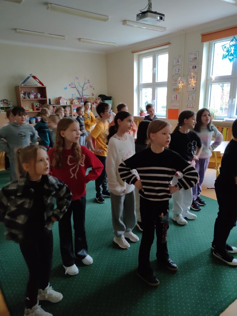 Na zdjęciu widzimy grupę dzieci w sali lekcyjnej, ustawionych w szeregu, prawdopodobnie podczas aktywności ruchowej lub ćwiczeń. Dzieci mają różnorodne stroje – kolorowe bluzy, swetry i wygodne buty. Sala jest jasna, z dużymi oknami, ozdobiona dekoracjami na ścianach, takimi jak drzewo z motylami i rysunki. W tle znajdują się półki z zabawkami i materiałami edukacyjnymi, co wskazuje, że jest to przestrzeń przeznaczona dla młodszych uczniów.