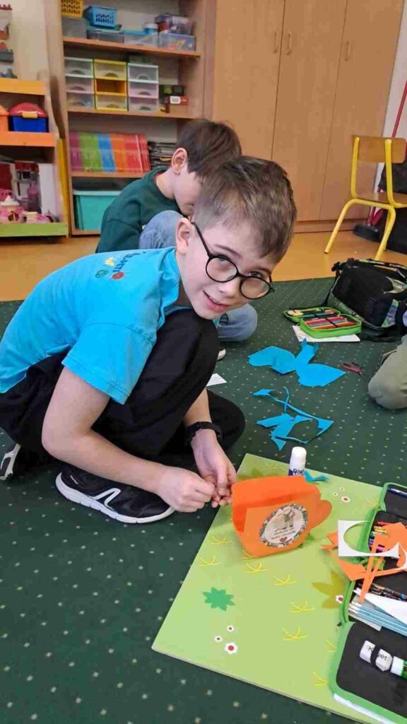 Na zdjęciu widać chłopca w okularach i niebieskiej koszulce kucającego na zielonym dywanie w ręku trzyma klej. Przed nim leży podkładka , na podkładce leży piórnik i wycięta pomarańczowa filiżanka z papieru. W tle widać drewniane szafki i półki z książkami oraz grami edukacyjnymi.