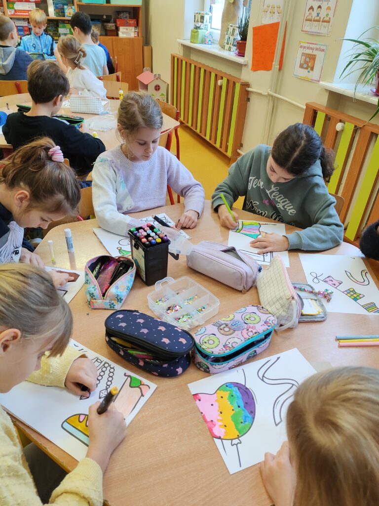 Na zdjęciu widać grupę dzieci w klasie podczas zajęć plastycznych. Siedzą przy stołach, skupione na kolorowaniu rysunków przedstawiających różne kształty i obiekty, np. lody i balony. Na stołach znajdują się przybory szkolne, takie jak piórniki, kredki, mazaki oraz pudełko z koralikami. W tle widać inne dzieci, półki z grami oraz kolorowe dekoracje na ścianach klasy