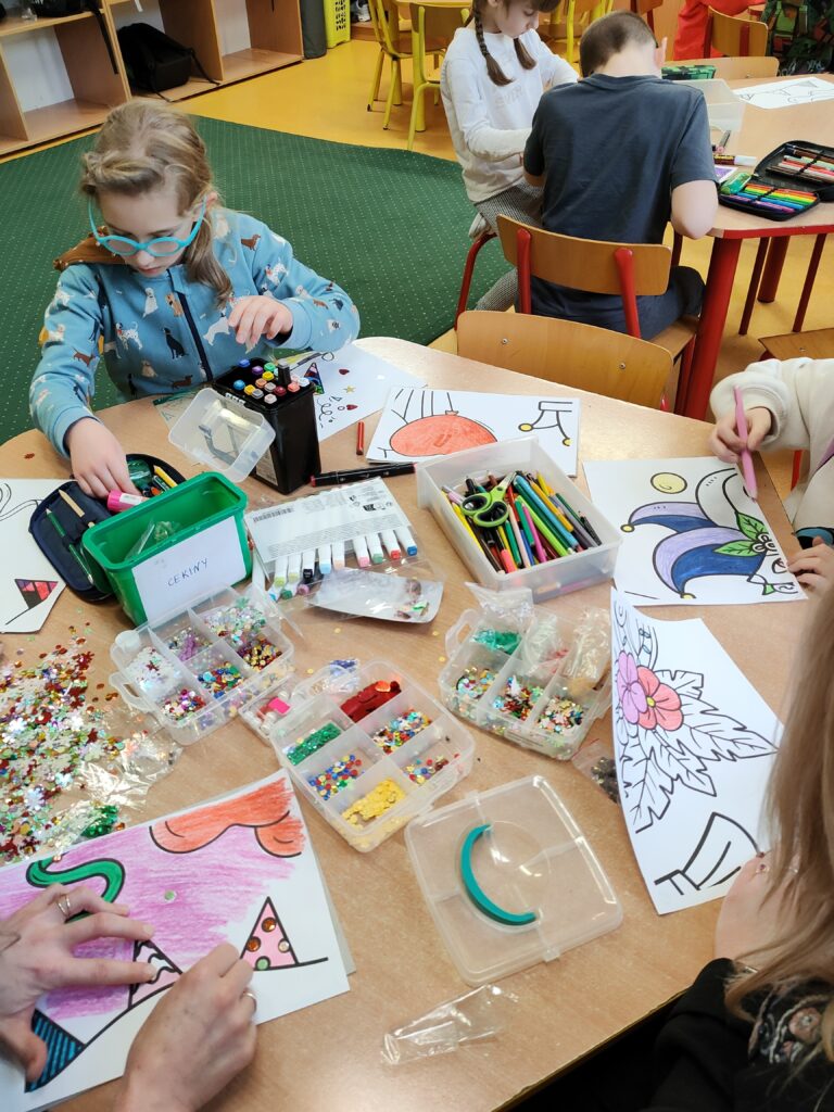 Na zdjęciu dzieci pracują nad kolorowaniem i ozdabianiem rysunków w klasie. Na stole znajduje się wiele przyborów plastycznych, takich jak kredki, mazaki, nożyczki, brokat oraz pojemniki z cekinami i innymi dekoracjami. Jedno z dzieci skupia się na wyborze materiałów, podczas gdy inne precyzyjnie kolorują swoje prace. W tle widać dalszą część klasy, z półkami i innymi uczniami zajętymi podobnymi aktywnościami.