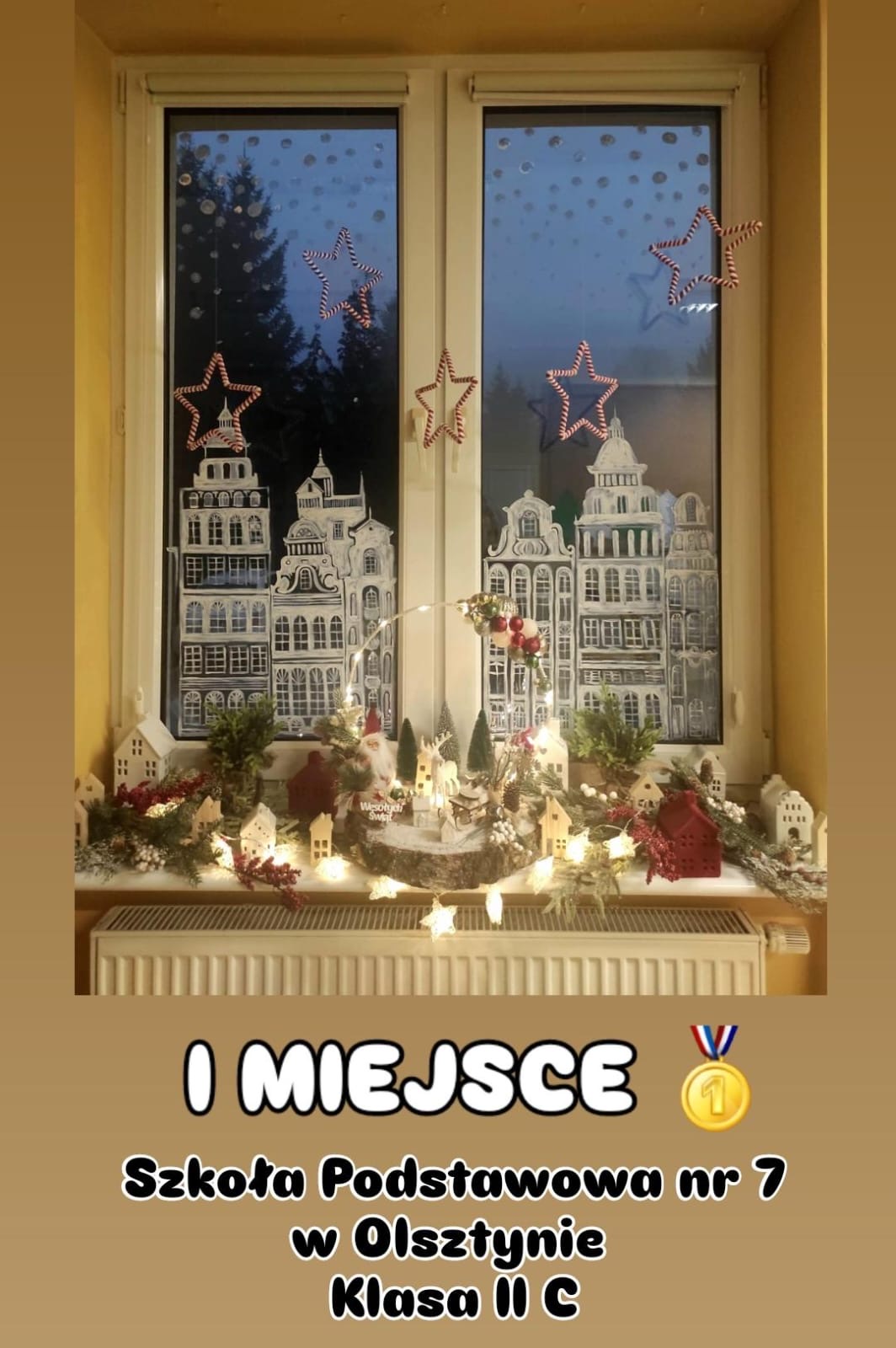 Na zdjęciu widzimy pięknie udekorowane okno, które zdobyło pierwsze miejsce w konkursie. Na szybach namalowane są białe budynki w stylu kamienic, przypominające świąteczny pejzaż miasta. Dodatkowo na szybach widać małe kropki imitujące śnieg oraz ozdobne gwiazdy wykonane z materiału w biało-czerwone paski. Na parapecie ustawiono świąteczne dekoracje: małe domki, światełka, gałązki z igliwiem, czerwone owoce oraz figurkę Mikołaja na drewnianym podłożu z napisem "Wesołych Świąt". Całość tworzy ciepły i przytulny klimat świąteczny. Na dole zdjęcia znajduje się napis informujący, że klasa II C ze Szkoły Podstawowej nr 7 w Olsztynie zdobyła pierwsze miejsce.