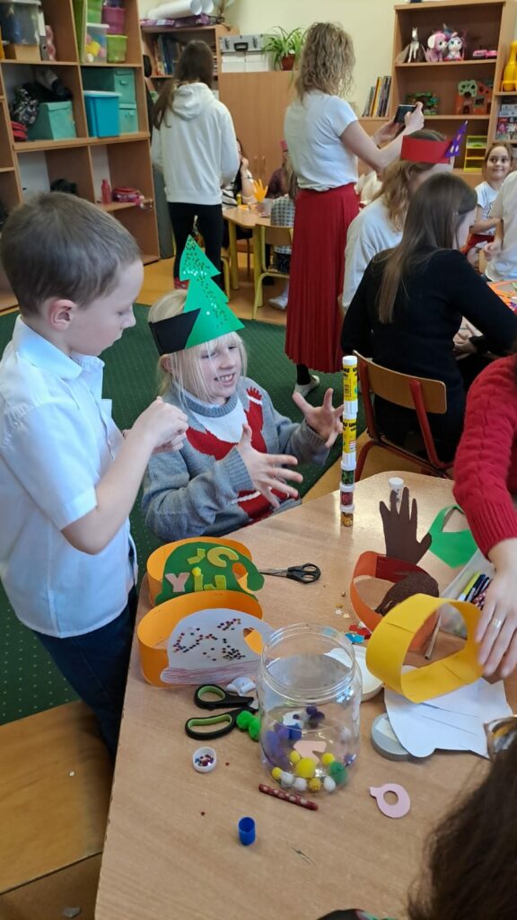 Na zdjęciu widzimy 2 dzieci (chłopcy)podczas zajęć plastycznych w świetlicy, które odbywają się w przedświątecznej atmosferze. Uczestnicy siedzą przy stole, tworząc kolorowe ozdoby, takie jak z wycinankami w kształcie choinki. Na stole znajdują się materiały plastyczne, takie jak papier kolorowy, klej i nożyczki, a na twarzach dzieci widoczne są uśmiechy, świadczące o dobrej zabawie.