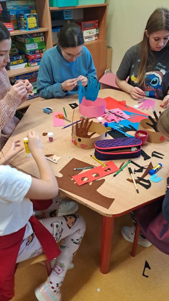 Na zdjęciu widzimy 3 studentki podczas zajęć plastycznych w świetlicy, które odbywają się w przedświątecznej atmosferze. Uczestnicy siedzą przy stole, tworząc kolorowe ozdoby, takie jak z wycinankami w kształcie dłoni. Na stole znajdują się materiały plastyczne, takie jak papier kolorowy, klej i nożyczki, a na twarzach dzieci i studentek widoczne są uśmiechy, świadczące o dobrej zabawie.