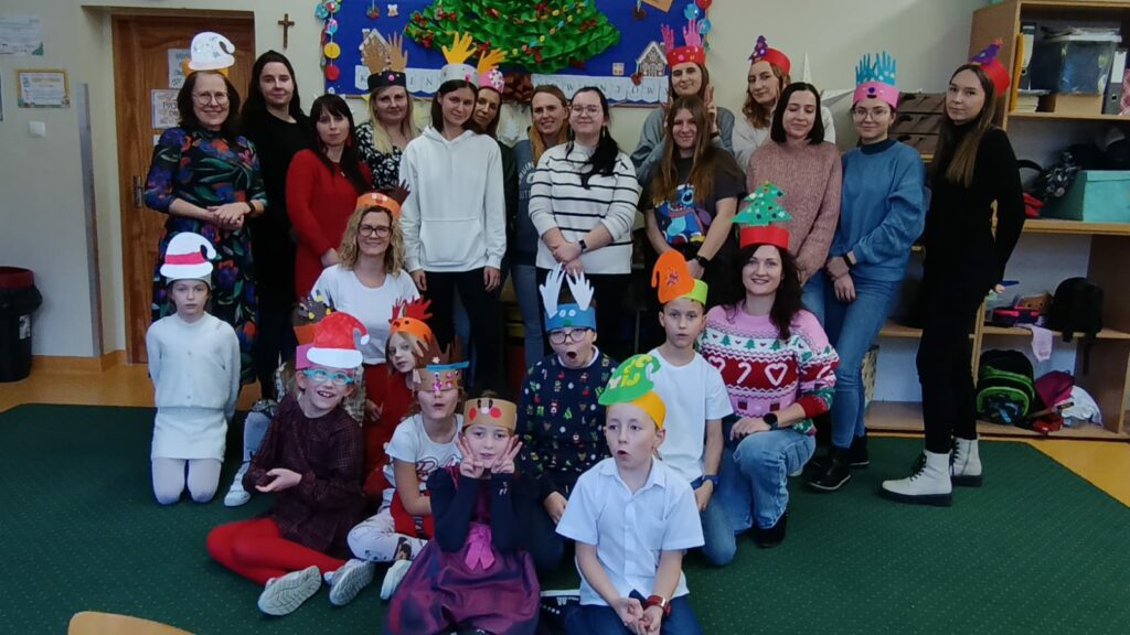 Na zdjęciu widzimy grupę dzieci oraz 13 studentek podczas zdjęcia wspólnego w świetlicy, które odbywają się w przedświątecznej atmosferze. Uczestnicy się są uśmiechy, co świadczy o dobrej zabawie. W tle jest tablica. Na tablicy choinka zrobiona z bibuły.