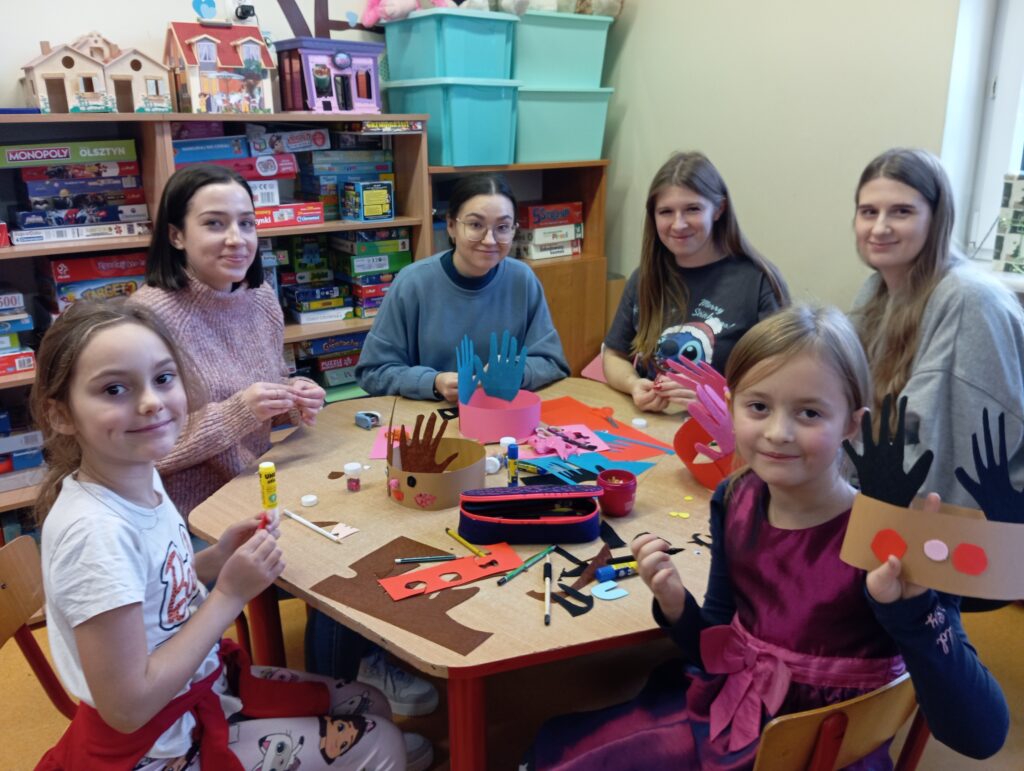 Na zdjęciu widzimy 2 dzieci (dziewczynki) i 4 studentki podczas zajęć plastycznych w świetlicy, które odbywają się w przedświątecznej atmosferze. Uczestnicy siedzą przy stole, tworząc kolorowe ozdoby, takie jak z wycinankami w kształcie dłoni. Na stole znajdują się materiały plastyczne, takie jak papier kolorowy, klej i nożyczki, a na twarzach dzieci i studentek widoczne są uśmiechy, świadczące o dobrej zabawie.