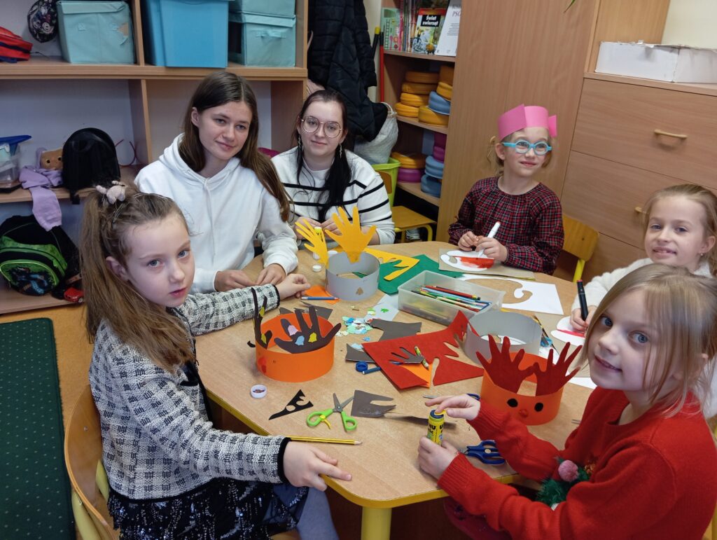 Na zdjęciu widzimy 4 dzieci (dziewczynki) i 2 studentki podczas zajęć plastycznych w świetlicy, które odbywają się w przedświątecznej atmosferze. Uczestnicy siedzą przy stole, tworząc kolorowe ozdoby, takie jak z wycinankami w kształcie dłoni. Na stole znajdują się materiały plastyczne, takie jak papier kolorowy, klej i nożyczki, a na twarzach dzieci i studentek widoczne są uśmiechy, świadczące o dobrej zabawie.