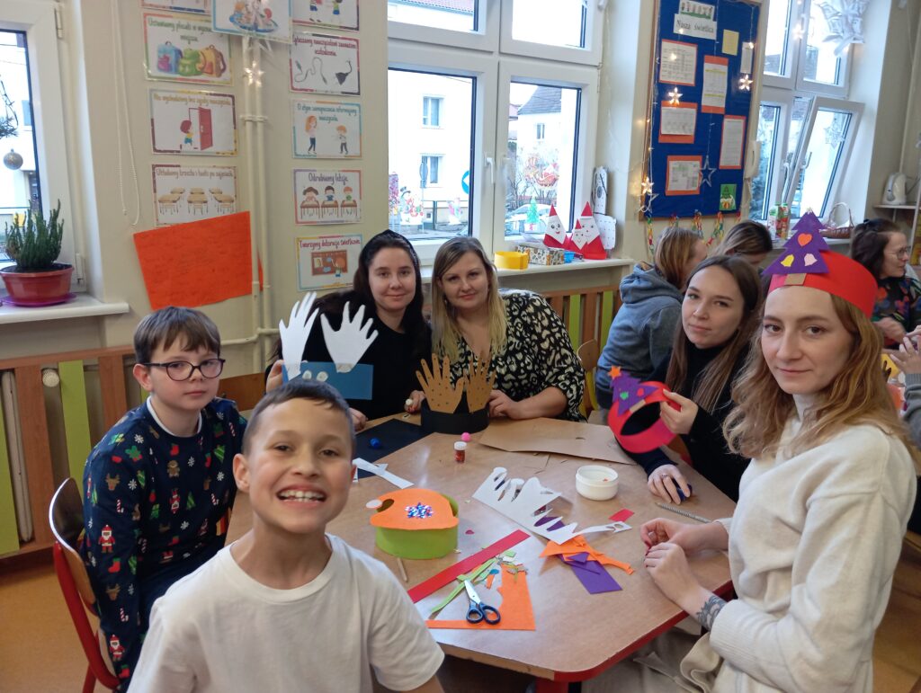 Na zdjęciu widzimy 2 dzieci (chłopców) i 4 studentki podczas zajęć plastycznych w świetlicy, które odbywają się w przedświątecznej atmosferze. Uczestnicy siedzą przy stole, tworząc kolorowe ozdoby, takie jak z wycinankami w kształcie dłoni. Na stole znajdują się materiały plastyczne, takie jak papier kolorowy, klej i nożyczki, a na twarzach dzieci i studentek widoczne są uśmiechy, świadczące o dobrej zabawie.