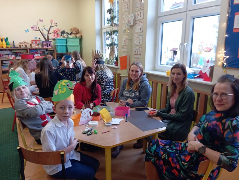 Na zdjęciu widzimy 2 dzieci (chłopców) i 3 studentki oraz 1 Nauczycielkę podczas zajęć plastycznych w świetlicy, które odbywają się w przedświątecznej atmosferze. Uczestnicy siedzą przy stole, tworząc kolorowe ozdoby, takie jak z wycinankami w kształcie dłoni. Na stole znajdują się materiały plastyczne, takie jak papier kolorowy, klej i nożyczki, a na twarzach dzieci i studentek widoczne są uśmiechy, świadczące o dobrej zabawie.