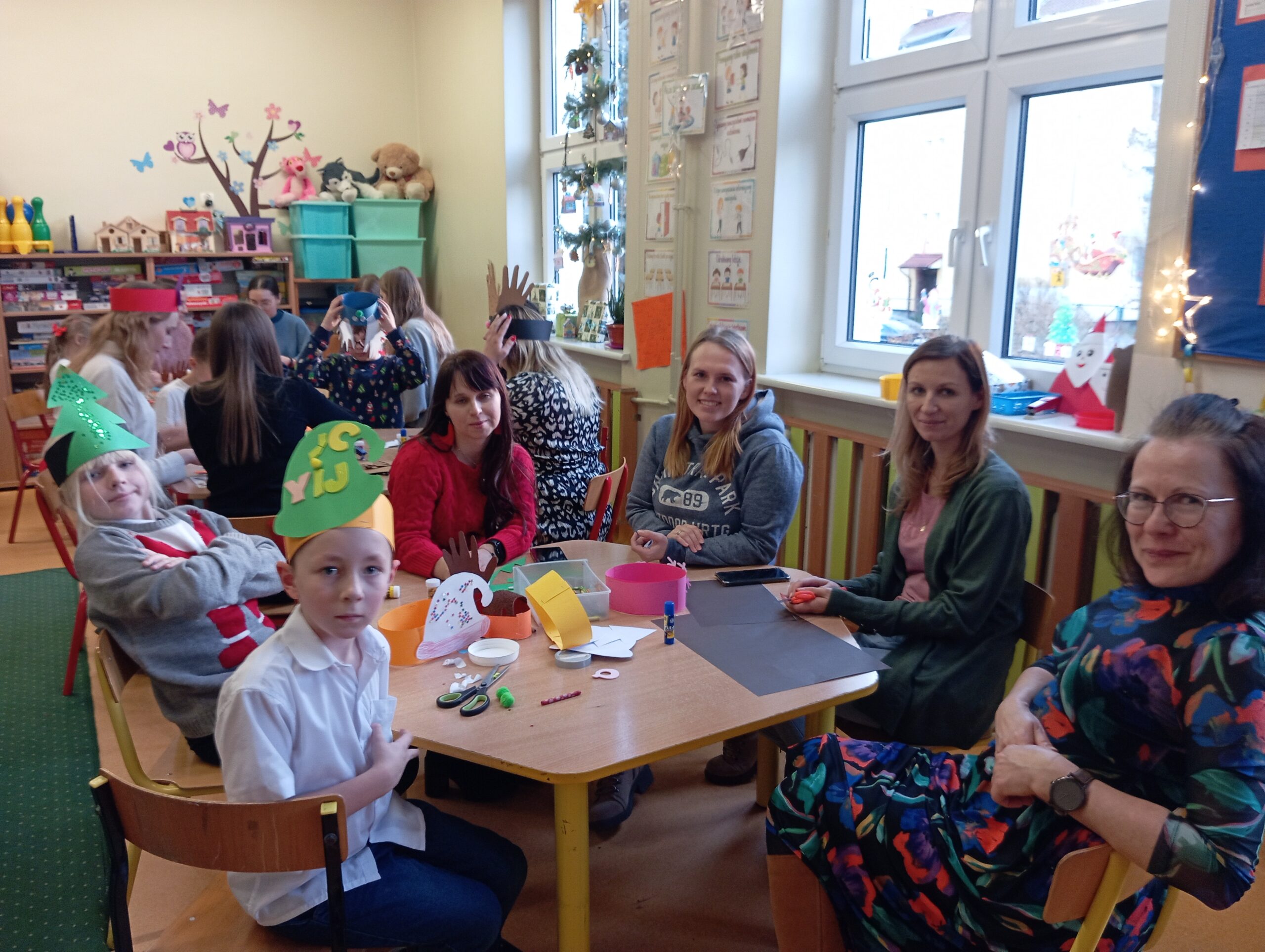 Na zdjęciu widzimy 2 dzieci (chłopców) i 3 studentki oraz 1 Nauczycielkę podczas zajęć plastycznych w świetlicy, które odbywają się w przedświątecznej atmosferze. Uczestnicy siedzą przy stole, tworząc kolorowe ozdoby, takie jak z wycinankami w kształcie dłoni. Na stole znajdują się materiały plastyczne, takie jak papier kolorowy, klej i nożyczki, a na twarzach dzieci i studentek widoczne są uśmiechy, świadczące o dobrej zabawie.