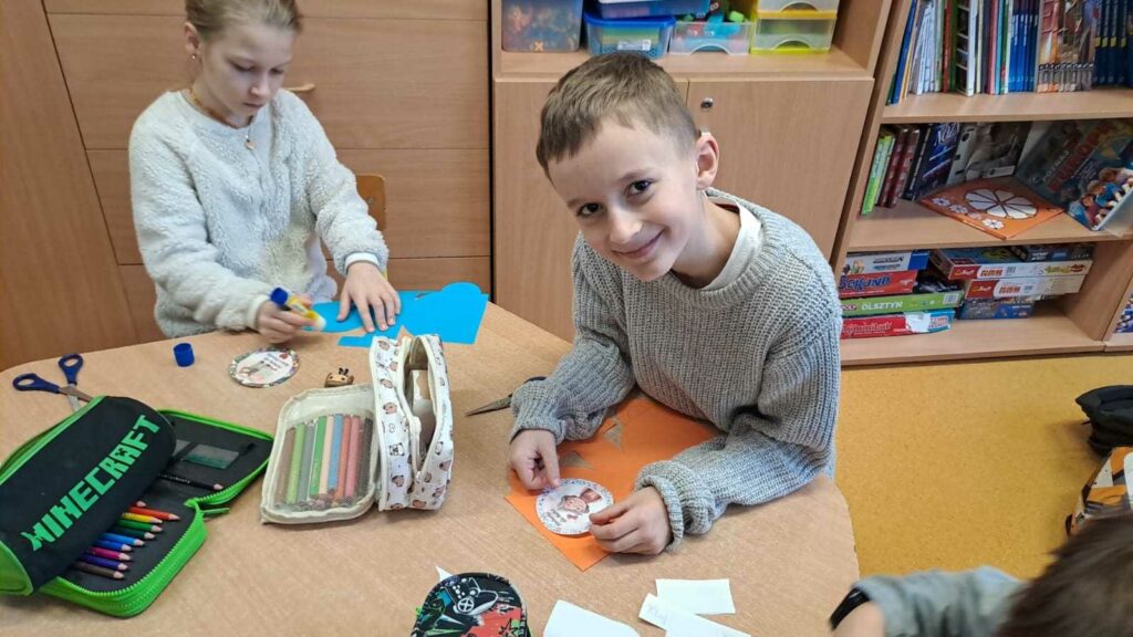 Na zdjęciu widać dwoje dzieci siedzących przy stole w klasie i wykonuje prace plastyczne, wycinając i przyklejając elementy na kolorowe kartki. Chłopiec na pierwszym planie uśmiecha się, dziewczynka siedząca obok jest skupiona na przyklejaniu wyciętych kształtów. Na stole znajdują się różne przybory szkolne, takie jak kredki, klej, nożyczki oraz piórniki, a w tle widać drewniane szafki i półki z książkami oraz grami edukacyjnymi.