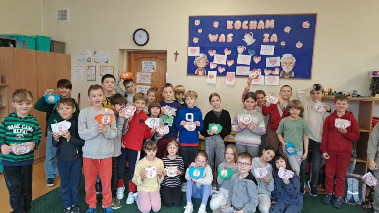 Na zdjęciu widać dużą grupę dzieci stojących w klasie i trzymających w rękach własnoręcznie wykonane prace plastyczne w kształcie filiżanek. W tle znajduje się niebieska tablica z napisem „KOCHAM WAS ZA” oraz rysunkiem starszej osoby, otoczona serduszkami i kartkami z napisami. Dzieci ubrane są w kolorowe stroje i wyglądają na zadowolone, prezentując swoje prace. Klasa jest wyposażona w drewniane szafki, półki z materiałami oraz zegar wiszący nad drzwiami. Atmosfera na zdjęciu jest ciepła i radosna, co sugeruje, że uczniowie uczestniczyli w specjalnej lekcji, być może związanej z Dniem Babci i Dziadka.