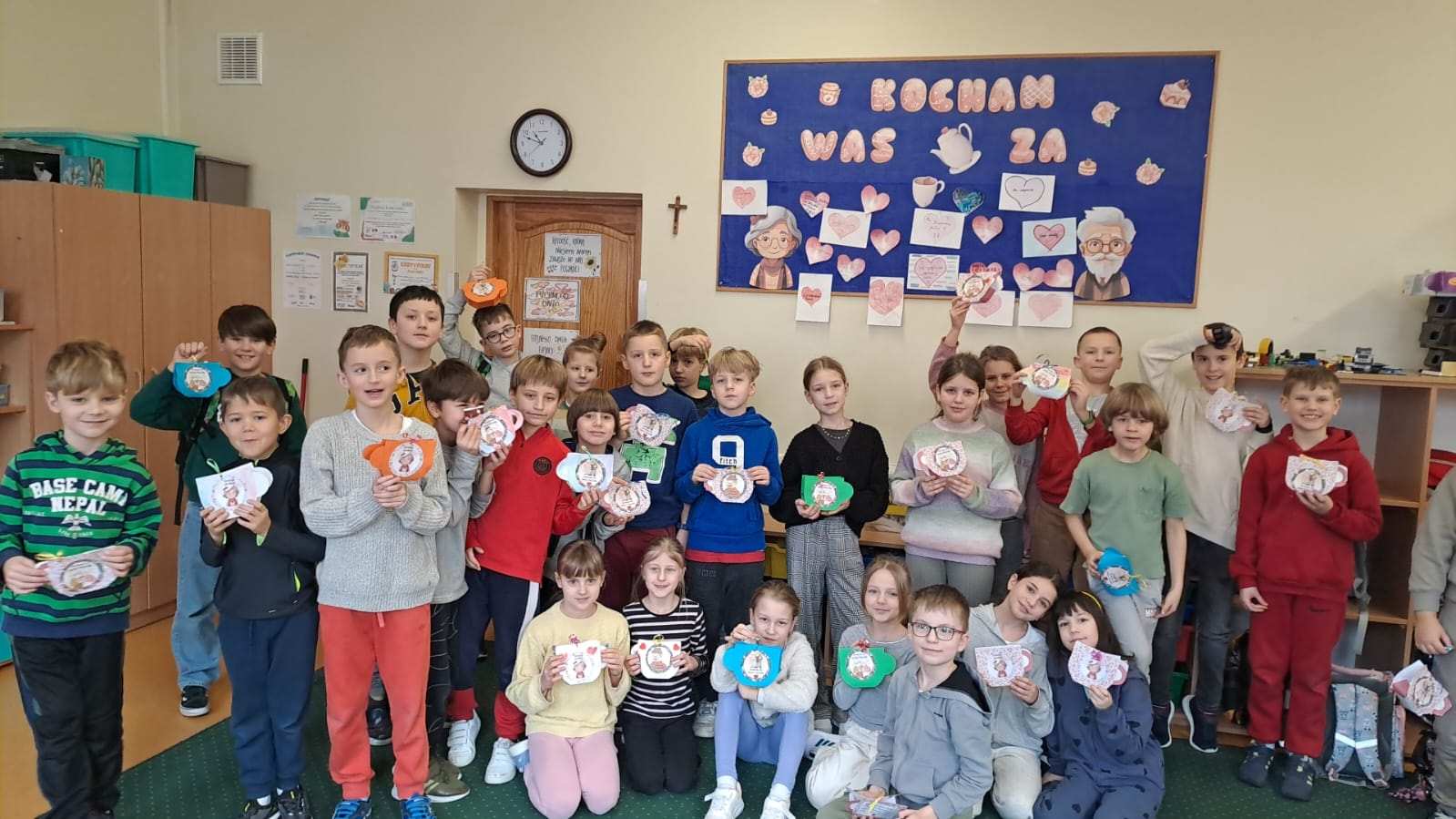 Na zdjęciu widać dużą grupę dzieci stojących w klasie i trzymających w rękach własnoręcznie wykonane prace plastyczne w kształcie filiżanek. W tle znajduje się niebieska tablica z napisem „KOCHAM WAS ZA” oraz rysunkiem starszej osoby, otoczona serduszkami i kartkami z napisami. Dzieci ubrane są w kolorowe stroje i wyglądają na zadowolone, prezentując swoje prace. Klasa jest wyposażona w drewniane szafki, półki z materiałami oraz zegar wiszący nad drzwiami. Atmosfera na zdjęciu jest ciepła i radosna, co sugeruje, że uczniowie uczestniczyli w specjalnej lekcji, być może związanej z Dniem Babci i Dziadka.