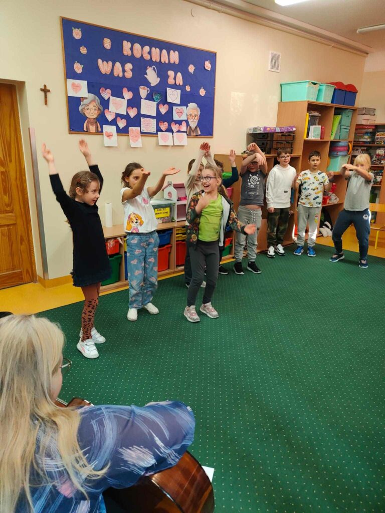 Na zdjęciu widać 8 dzieci będących w podskokach są na dywanie w kręgu. Na pierwszym planie widać siedzącą tyłem kobietę, która trzyma w ręku gitarę. Ze zdjęcia można wywnioskować, że jest to zabaw muzyczna.