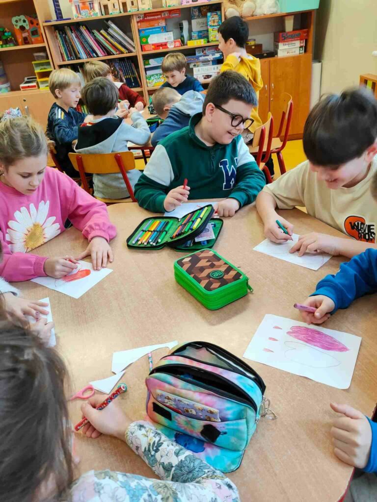 Na zdjęciu widać 5 dzieci siedzących przy stoliku 1 dziewczynka i 4 chłopców. Dzieci wykonują czerwone serca na białych papierze. Na stoliku widać 3 kolorowe piórniki. W tle widać stolik i siedzące przy nim dzieci 6 chłopców oraz regał z książkami.