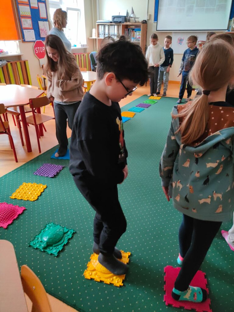 Na zdjęciu znajduje się 7 dzieci bawiących się w sali świetlicowej. Na pierwszym polanie widać chłopca, który ma okulary, czarną bluzę, ciemne włosy. Po lewej stronie widać dziewczynkę w brązowej bluzie z rozpuszczonymi włosami. Za dziewczynką stoi Pani w niebieskiej bluzce. W kolejnym planie widać obok siebie dziewczynkę w białej bluzce, chłopca z krótkimi włosami w ciemno niebieskiej bluzce, Obok jest chłopiec w okularach w bluzce z napisami. Dzieci chodzą po matach sensorycznych w skarpetkach, utrzymując równowagę . Jest to zabawa rozwijająca motorykę i zmysł dotyku, zmysł wzroku. W tle widać regały z książkami, zabawkami i pomocami dydaktycznymi, a także ekran projekcyjny, drukarkę.