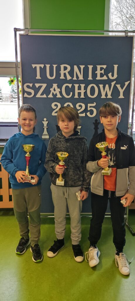 na zdjęciu widać 3 chłopców - zwycięzców Turnieju z pucharami w ręku. Za nimi tablica z napisem Turniej Szachowy 2025