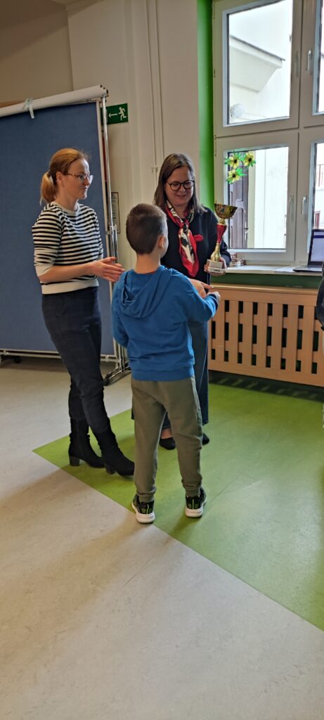 na zdjęciu widać chłopca stojącego tyłem. który odbiera puchar. Wręcza go pani dyrektor Dorota Zabuska w okularach z chustą na szyi w kolorze biało - czerwonym. Obok stoi druga pani w okularach, w bluzie w paski i granatowych spodniach.