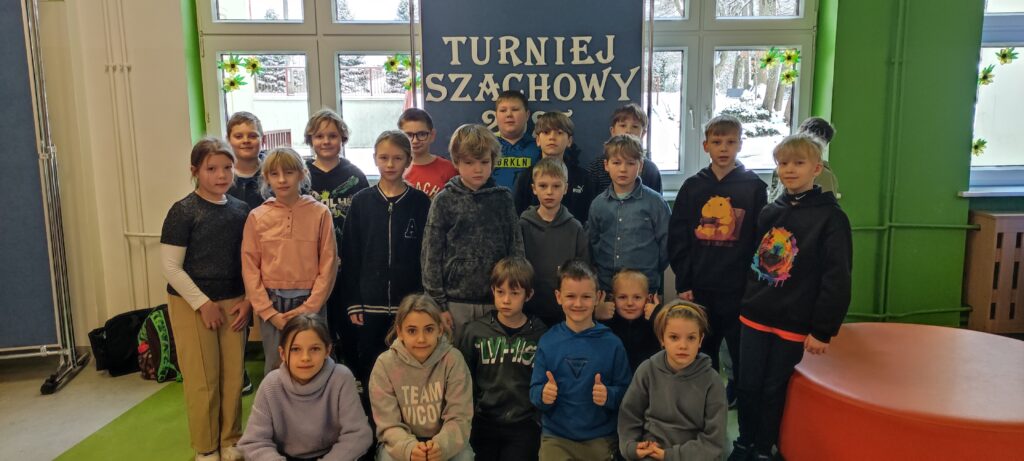 zdjęcie przedstawia grupę 21 dzieci - wśród nich kilkoro stojących i kilkoro kucających. Za nimi stojąca tablica a na niej biały napis Turniej Szachowy
