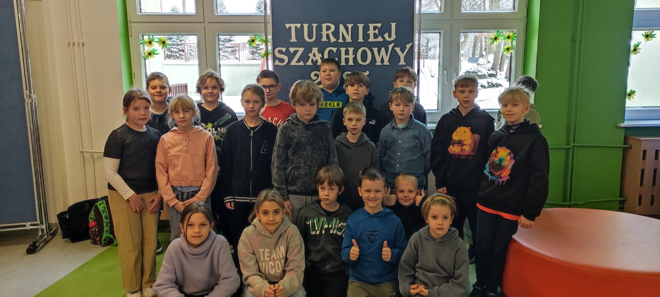 zdjęcie przedstawia grupę 21 dzieci - wśród nich kilkoro stojących i kilkoro kucających. Za nimi stojąca tablica a na niej biały napis Turniej Szachowy